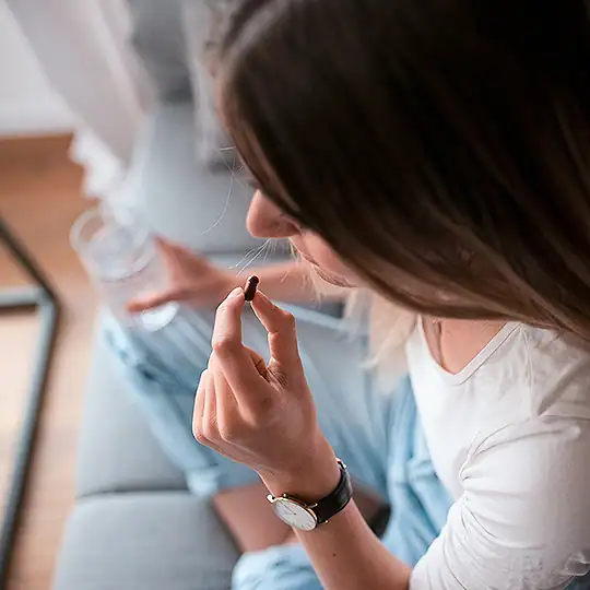 Kapsel rubyni Schwarze Johannisbeere – zur täglichen Anwendung Frau sitzt im Schneidersitz auf einem Sofa, hält eine rubyni Kapsel Schwarze Johannisbeere in der einen und ein Glas Wasser in der anderen Hand