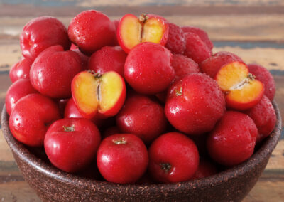 Auf einem Holztisch steht eine braune Schale voll mit Acerola-Kirschen. Drei Früchte sind aufgeschnitten und zeigen das gelbe Fruchtpulver.