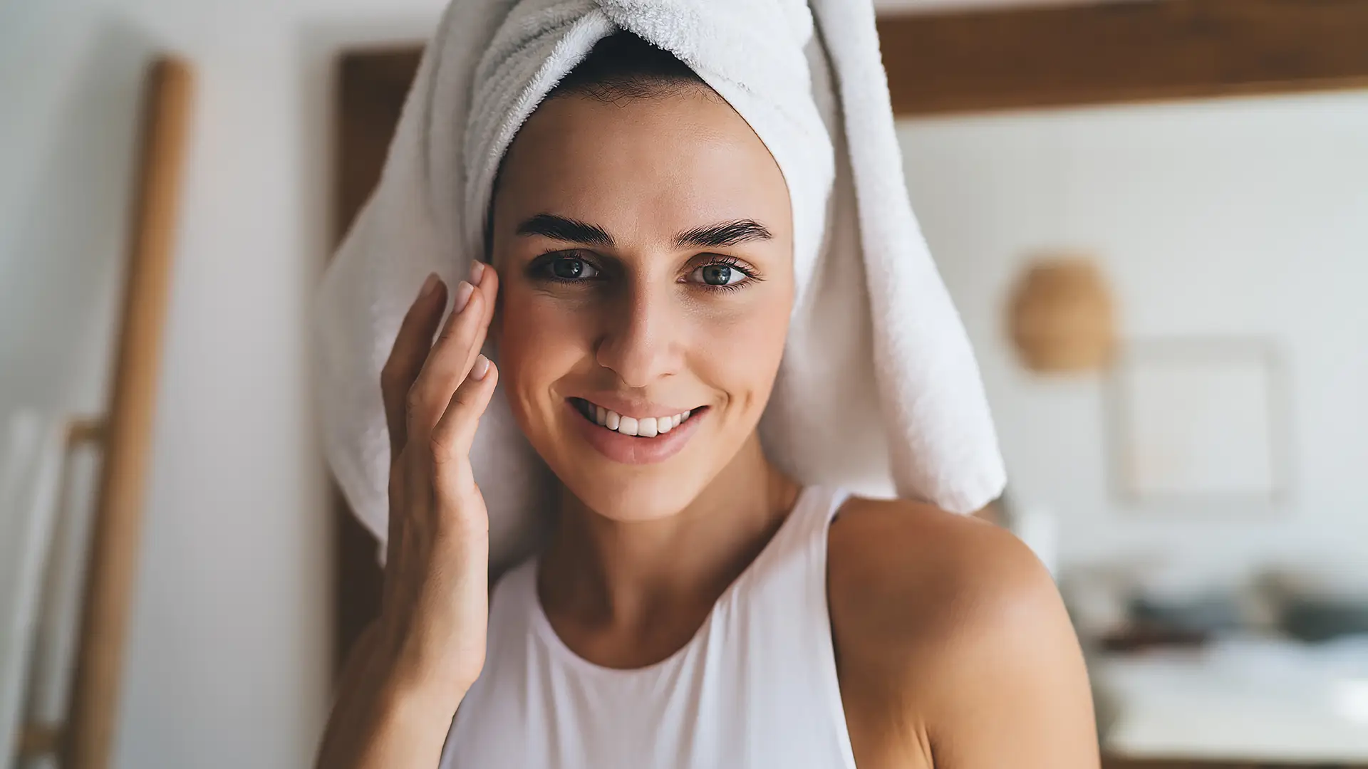 Junge Frau mit weißem Handtuchturban streicht sich sanft über die Wange – Symbol für natürliche Hautpflege, Antioxidans und Kollagenbildung.
