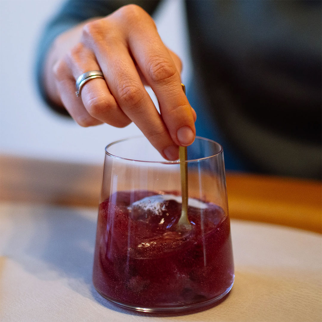 Ballaststoffe fürs Mikrobiom – trinkfertig in Sekunden Frau rührt mit einem Teelöffel in einem Glas Wasser, das sich durch das rubyni® MUCOSA Pulver tief dunkelrot verfärbt hat.